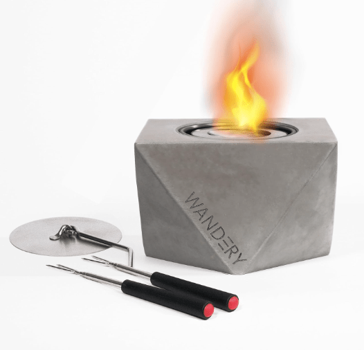 A portable tabletop fire pit featuring a geometric gray design, with flames visible at the top. The image includes roasting sticks and a protective cover.