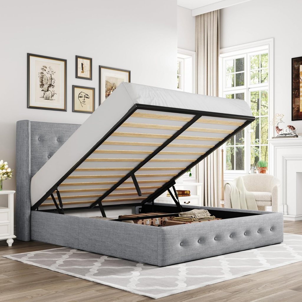 A light grey lift-up storage bed with a button-tufted headboard, partially raised to show the storage space underneath, set in a bright room with large windows and decorative wall art.
